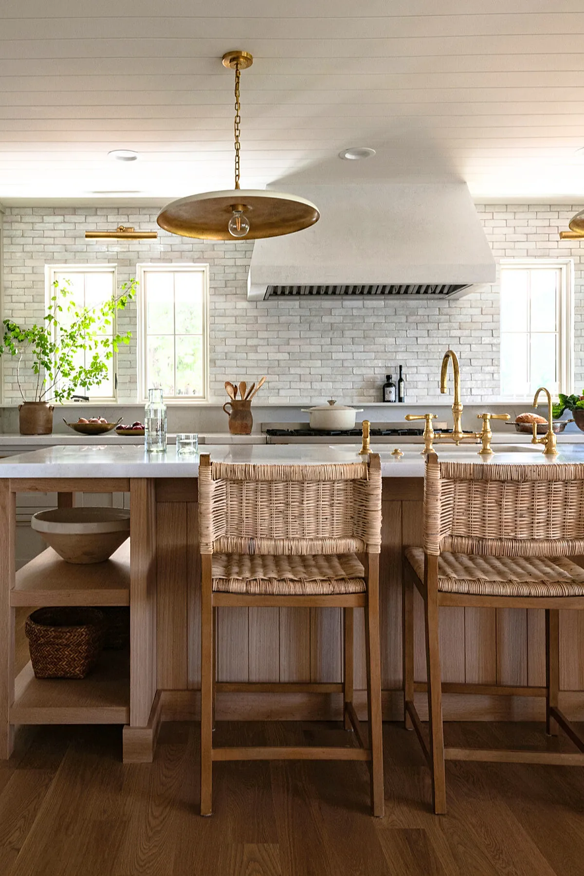 Bright kitchen featuring a white brick backsplash, wooden island with open shelving, woven bar stools, white countertop, brass faucet and pendant lighting, and two windows letting in natural light.