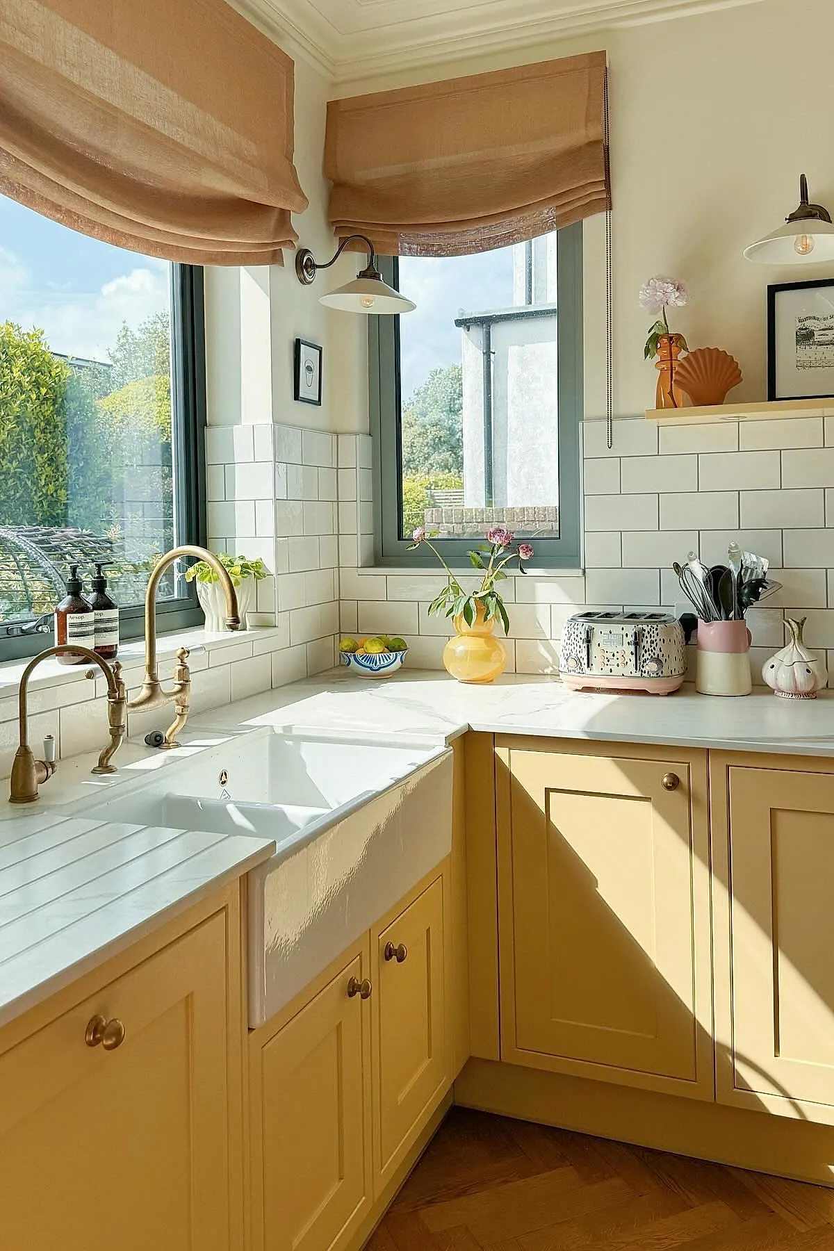 Yellow cottage kitchen with light yellow kitchen walls, white farmhouse sink featuring a vintage brass faucet, white subway tile backsplash, beige Roman shades on windows, yellow cabinetry below, a patterned toaster in pastel colors, and small decorative items including flowers and utensils on the marble countertop.