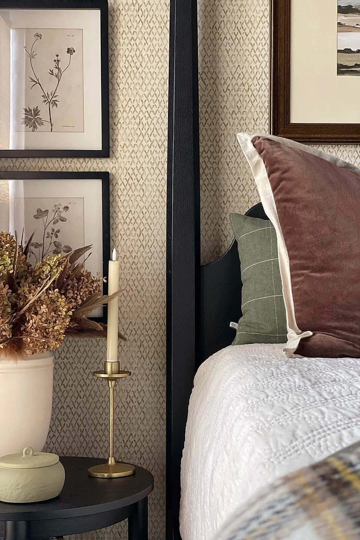 Close-up of an earthy bedroom corner with a black round side table holding a ceramic pot of dried florals, a brass candle holder, framed botanical prints, and layered neutral bedding