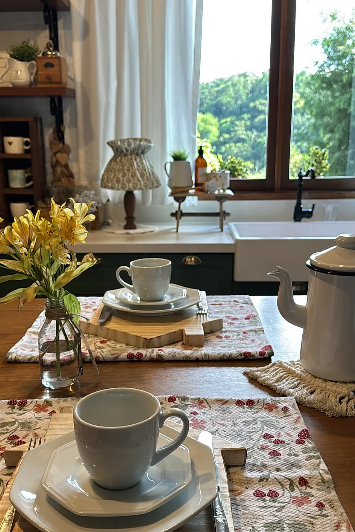 Cozy cottage kitchen table set with white ceramic cups and plates on layered wooden trays, floral patterned placemats, yellow flowers in a small glass vase, a patterned fabric lampshade on the counter, and large window letting in natural light revealing green foliage outside.