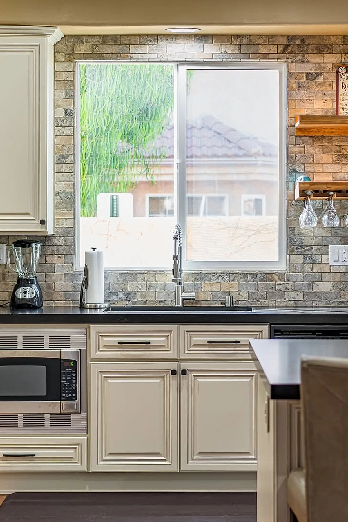 Kitchen featuring cream cabinetry with black handles, a multitoned brick backsplash surrounding a large window above the stainless steel sink with a modern faucet, black countertop, and wooden shelves holding glassware to the right.