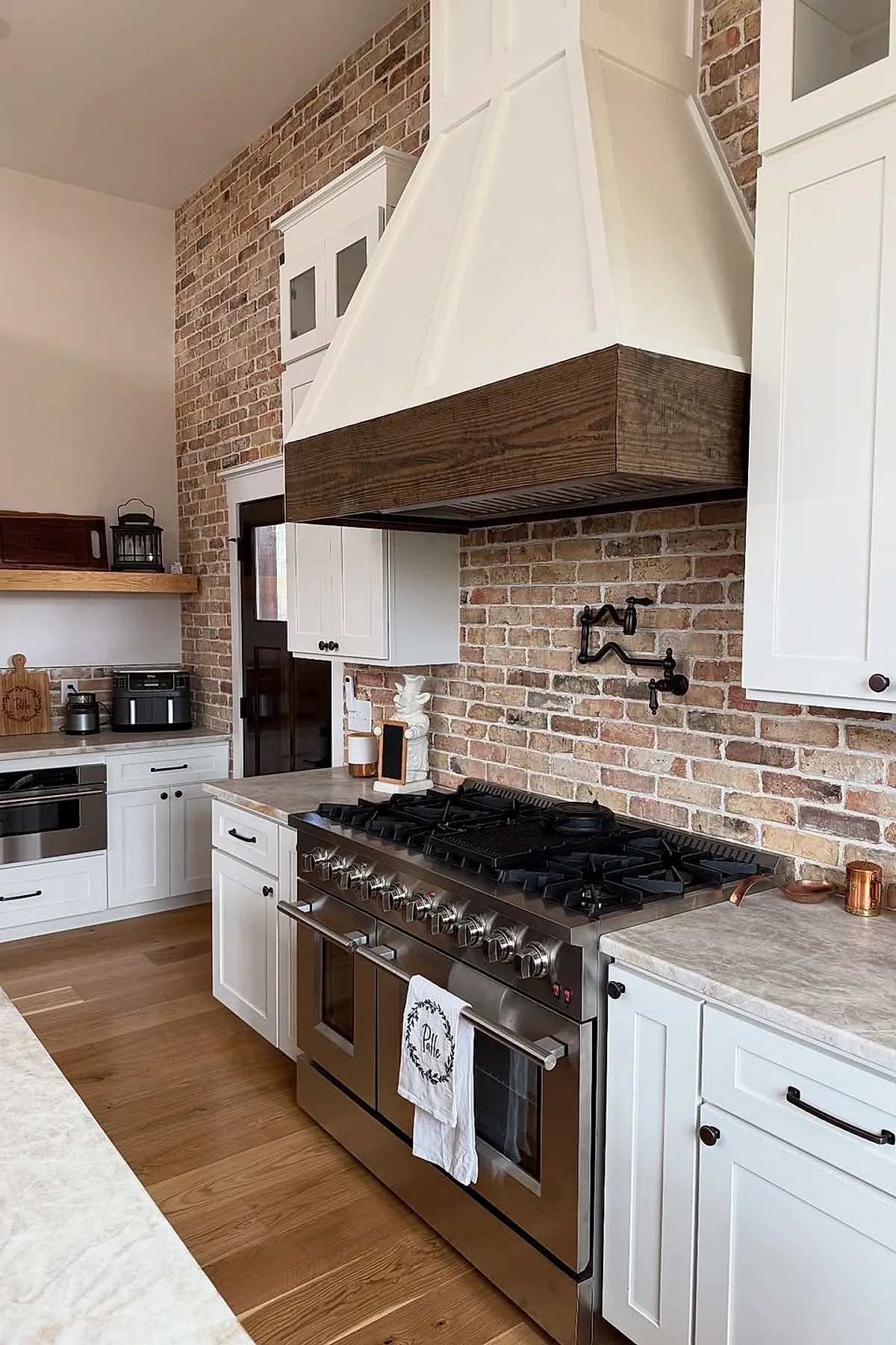 Kitchen featuring an exposed brick backsplash behind a large stainless steel gas range with multiple burners, white cabinetry, light gray countertops, wooden flooring, and a white range hood with dark wood trim.