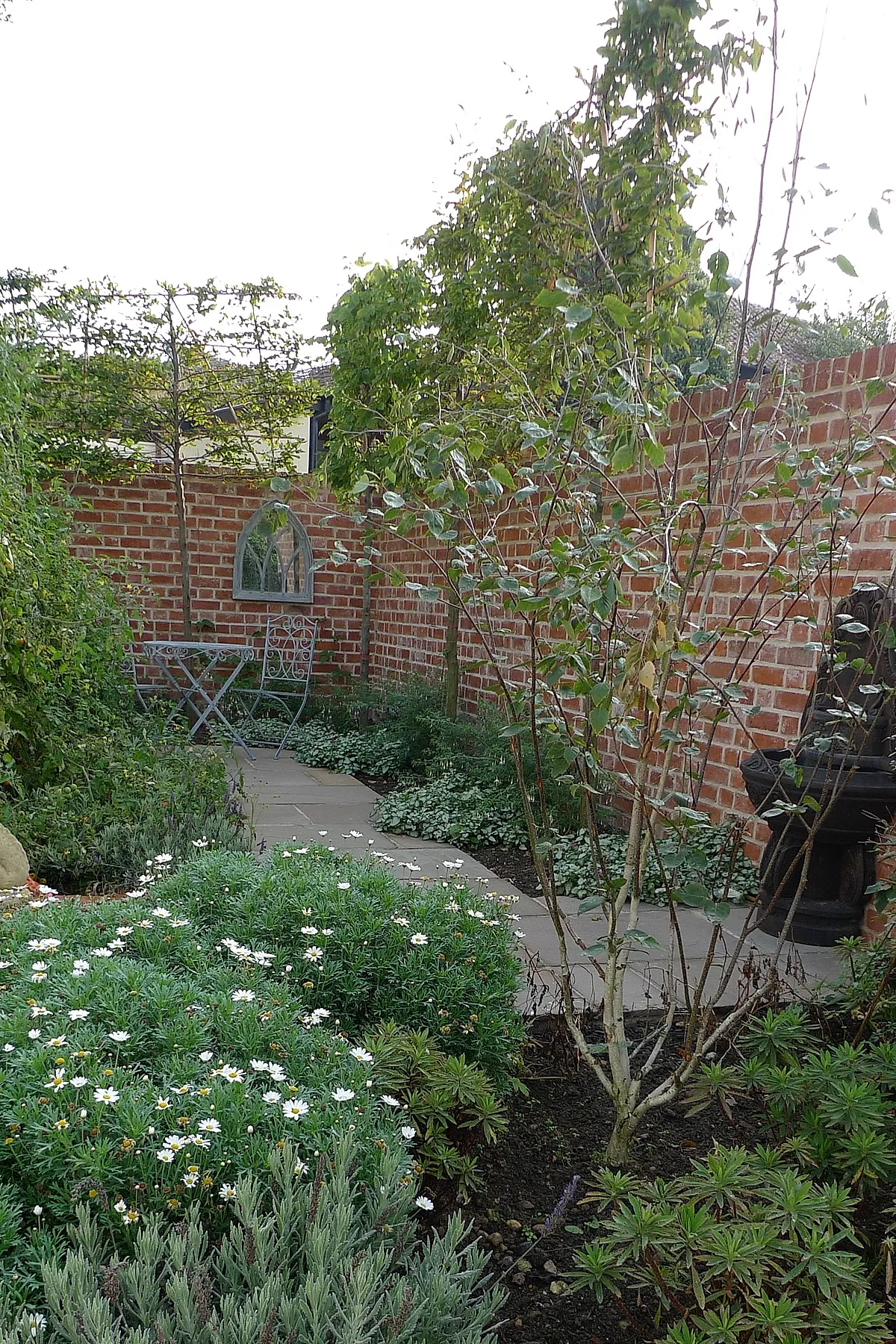 Small courtyard garden featuring red brick walls, narrow stone pathway, dense green plants and white daisies bordering the path, small round metal table with two chairs in muted blue, and climbing plants along the walls.