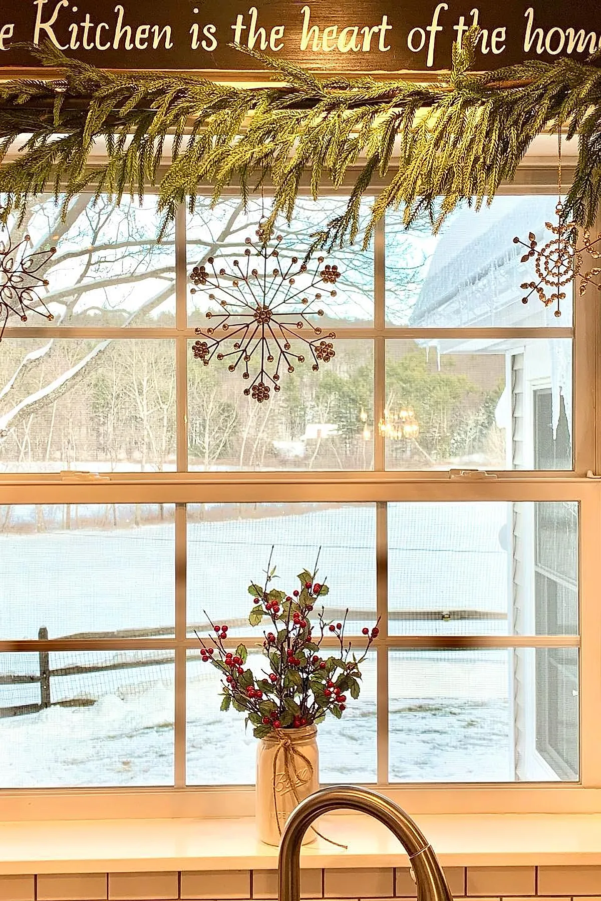 christmas window decorations featuring green pine garland draped above a white-framed kitchen window, with hanging wire snowflake ornaments and a mason jar vase filled with red berry branches sitting on the windowsill, all against a snowy outdoor backdrop.