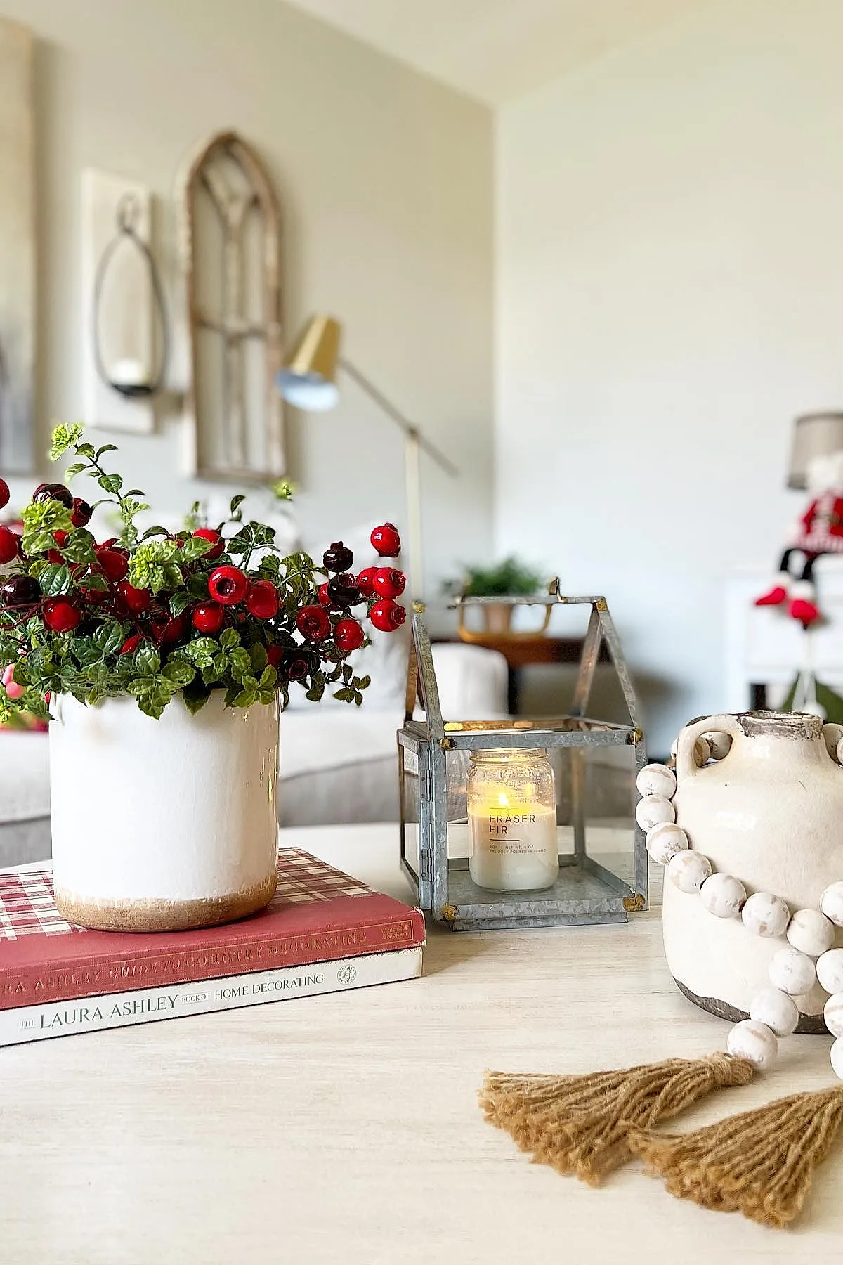 christmas coffee table decor featuring a white ceramic pot with green foliage and bright red berries, a lit candle inside a metal lantern, and an off-white ceramic vase adorned with chunky wooden bead garland with tassels, all arranged on a light wood coffee table with two hardcover books underneath the planter.