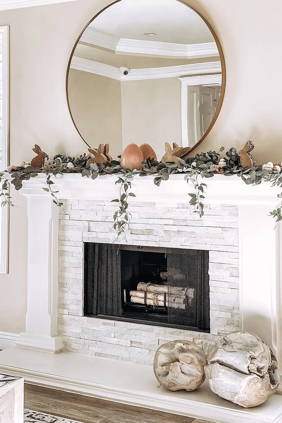 White stone fireplace mantel decorated with green eucalyptus garland, wooden and cork bunny figurines, pastel-colored eggs, and a large round mirror reflecting the ceiling details.