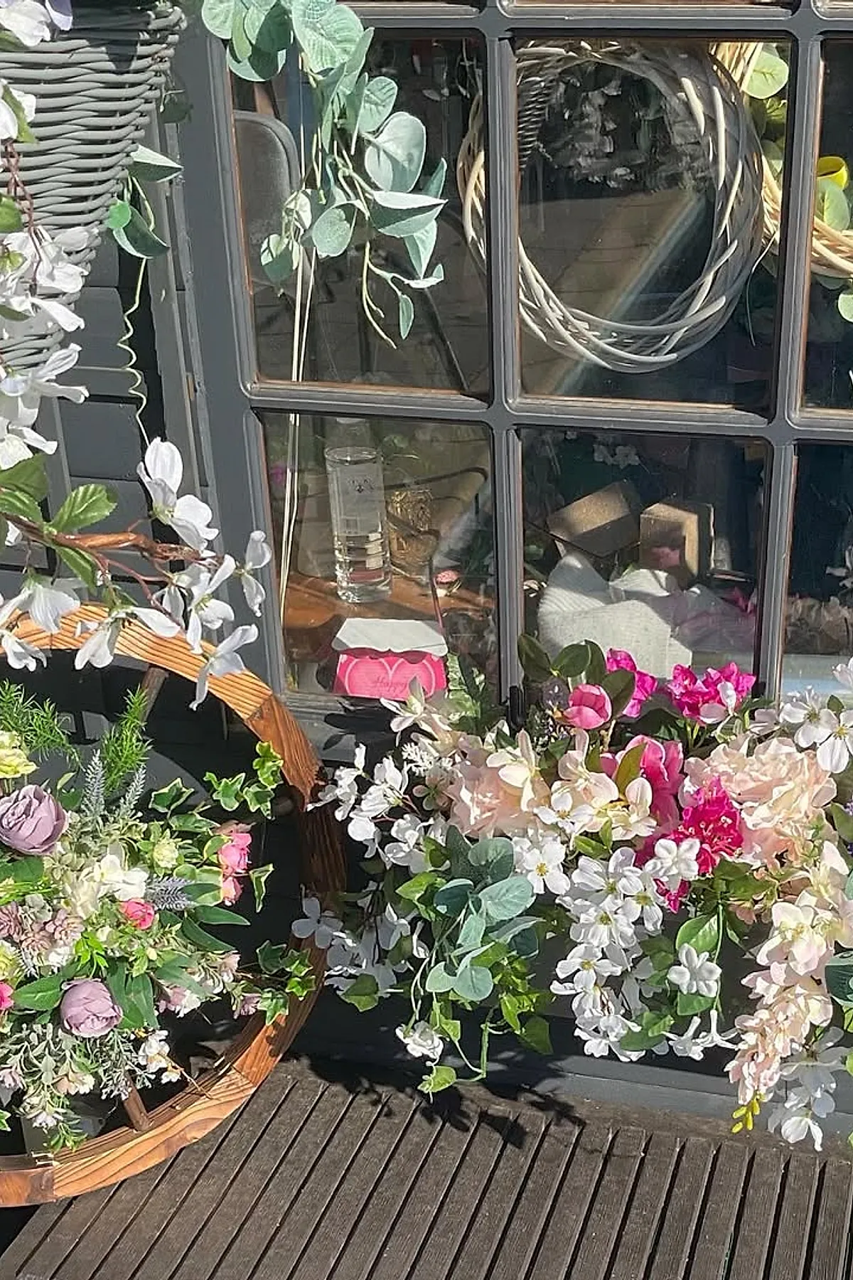 Colorful spring window box filled with bright pink, white, and soft blush flowers along with green leaves, hanging under dark-framed window on a wooden deck floor.