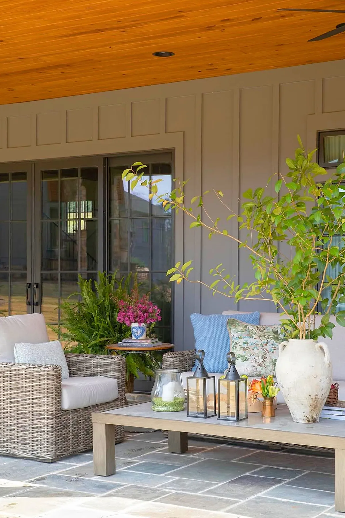 Covered patio attached to house featuring rattan armchair and sofa with cream cushions, wooden coffee table with decorative lanterns, large ceramic vase with greenery, potted fern and pink flowers on side table, gray paneled wall, and wooden ceiling.