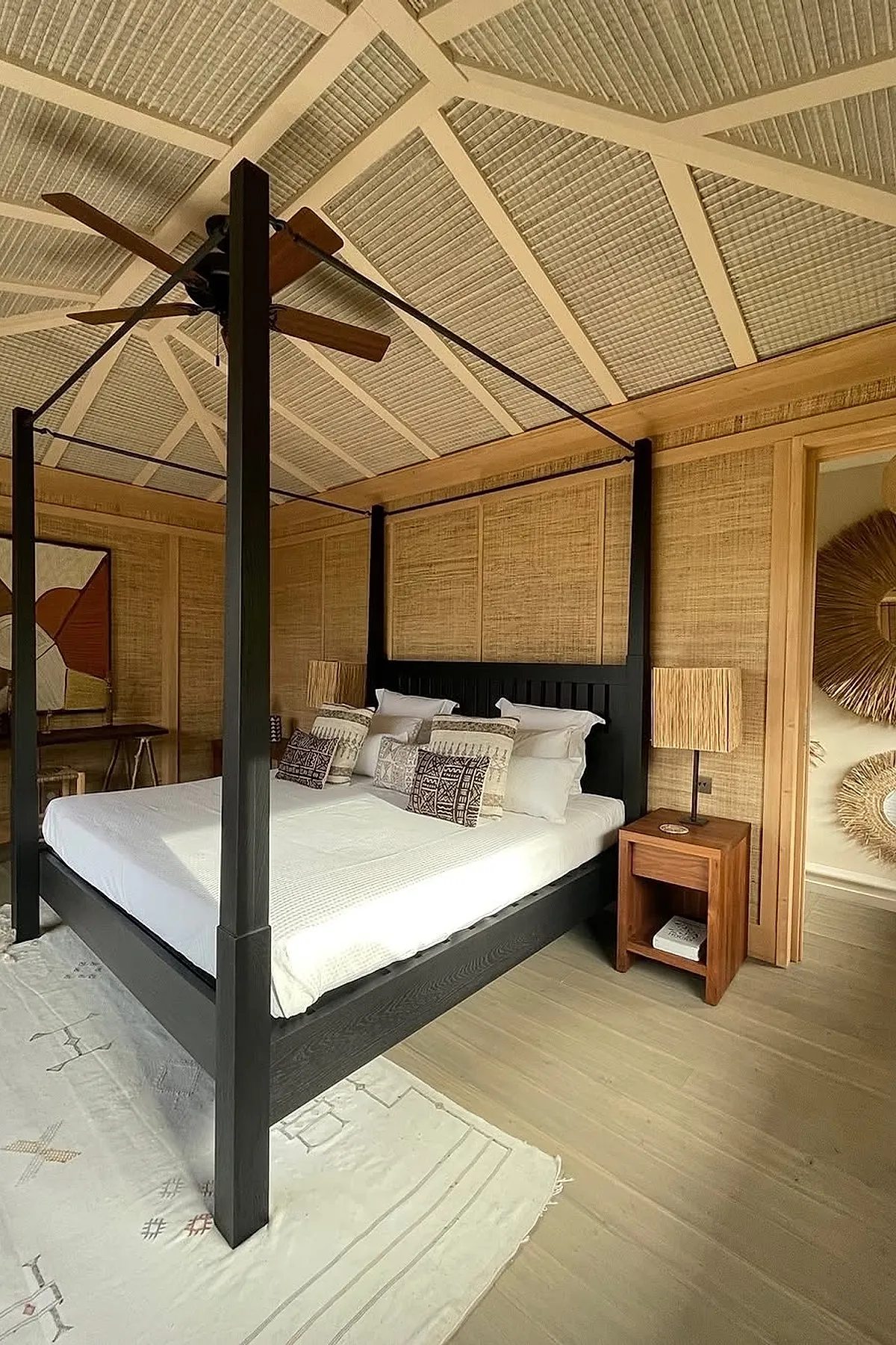 Modern bohemian bedroom with a black four-poster canopy bed dressed in white bedding and patterned throw pillows, natural woven wall panels, wooden nightstands with textured lamps, light wood flooring, and ceiling fan under a textured beige vaulted ceiling.