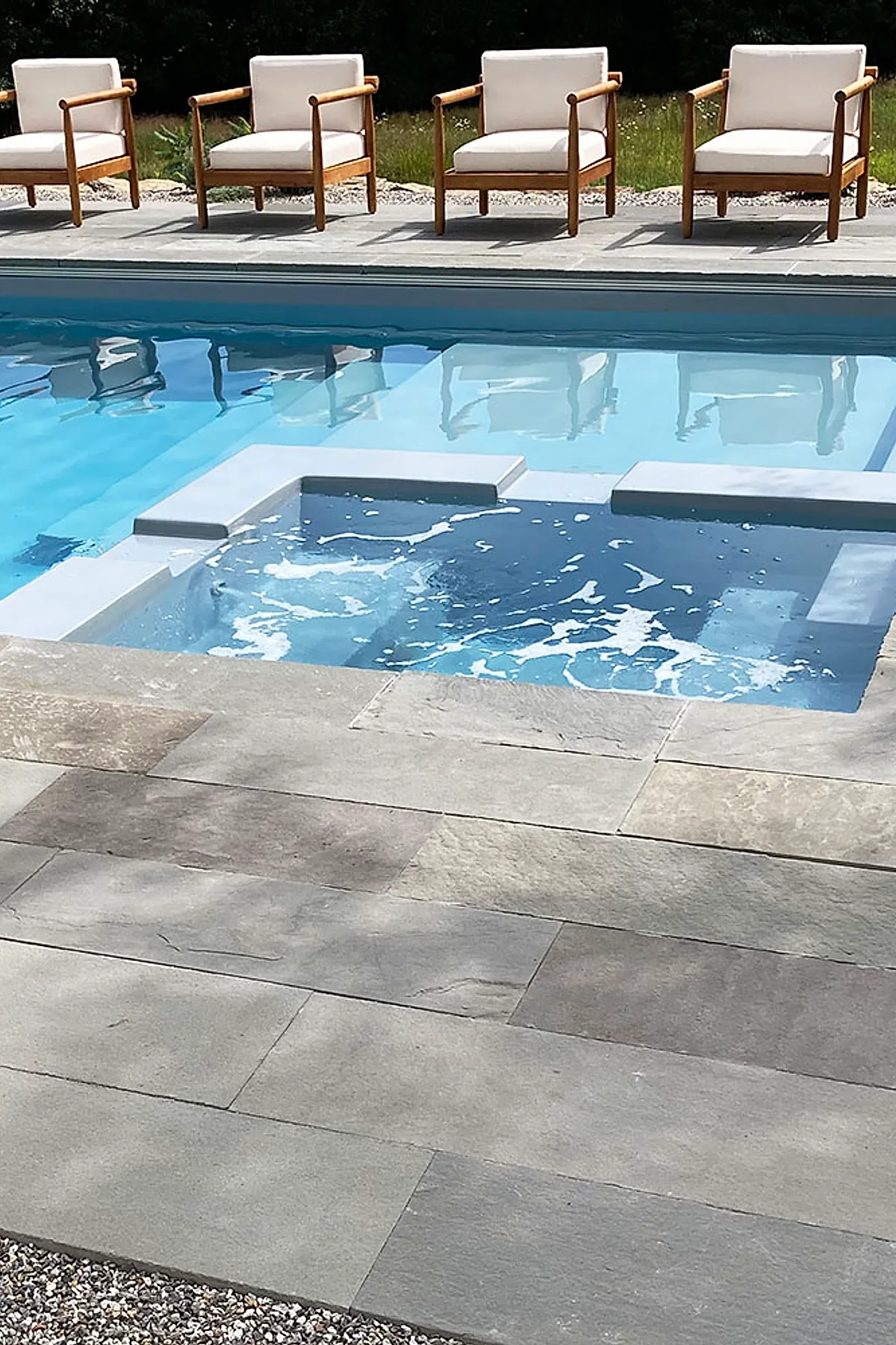 Modern pool deck with large rectangular neutral stone tiles surrounding a swimming pool, featuring five wooden lounge chairs with white cushions aligned along the far edge under bright daylight.