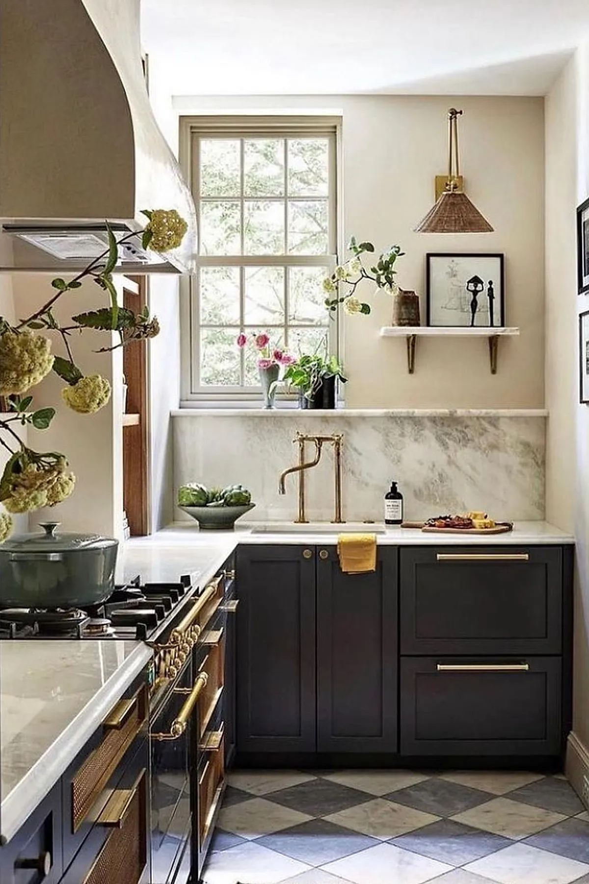 small kitchen with dark gray lower cabinets, white marble countertops, gold faucet and hardware, gas stove with brass details, window above sink, decorative plants and framed artwork