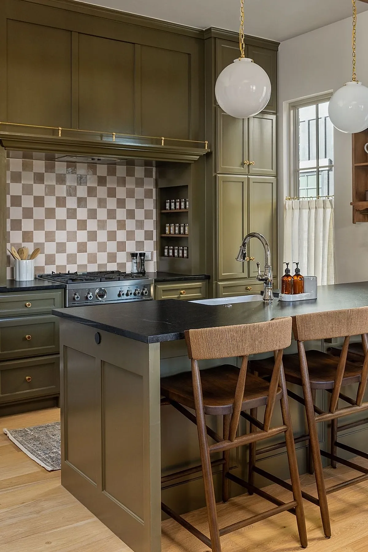 Cottage kitchen featuring a beige and light brown checkerboard tile backsplash behind a stainless steel stove, olive green cabinetry with brass knobs, black countertops, wooden bar stools, and large round white pendant lights hanging from the ceiling.