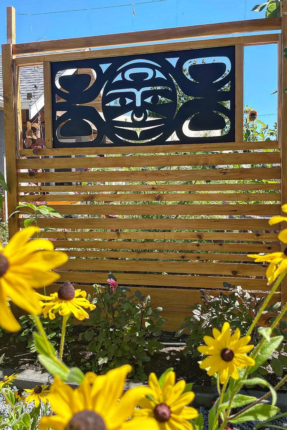 Wooden planter privacy wall with horizontal slats and a decorative black metal cutout panel above, surrounded by bright yellow flowers in a sunny outdoor garden