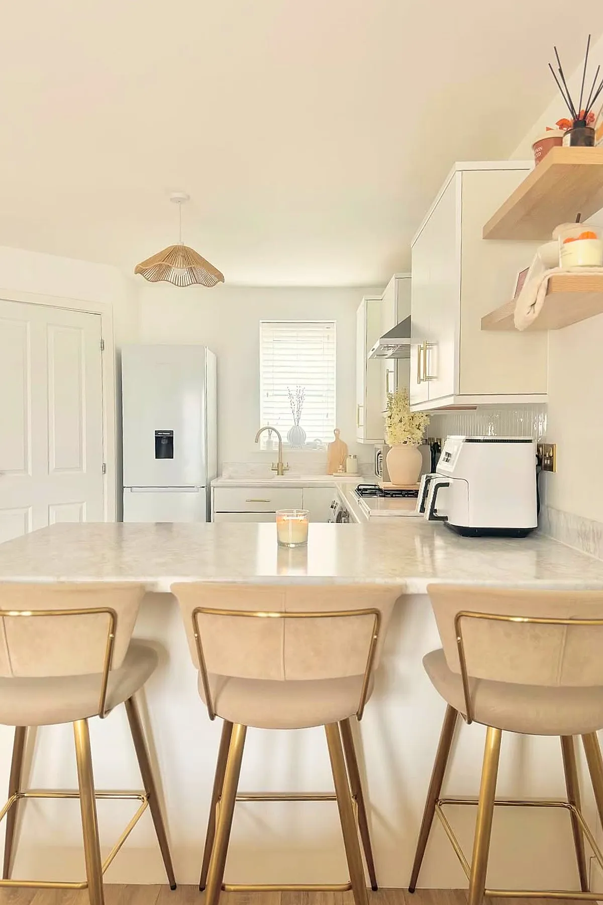 Warm neutral kitchen featuring cream marble countertop island with three beige bar stools with gold metal legs, white cabinets with gold handles, wooden floating shelves, and a rattan pendant light.