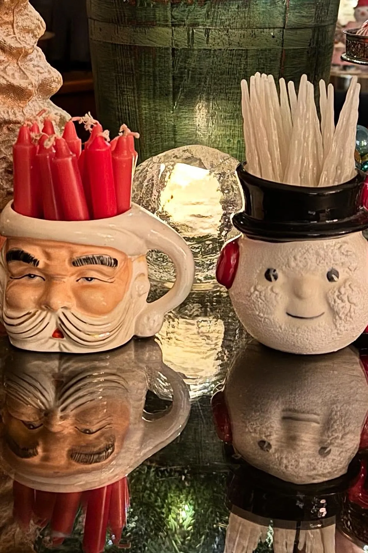 christmas coffee table decor featuring a ceramic Santa Claus mug holding red taper candles and a white snowman face container with a black top hat filled with white taper candles, set on a reflective surface with warm lighting