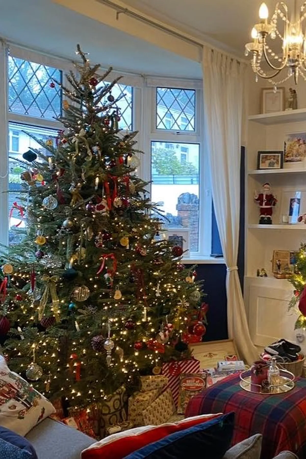 A tall Ralph Lauren Christmas tree decorated with warm white lights, red and gold ribbons, and traditional ornaments surrounded by wrapped gifts in patterned paper beside bay windows with white curtains.