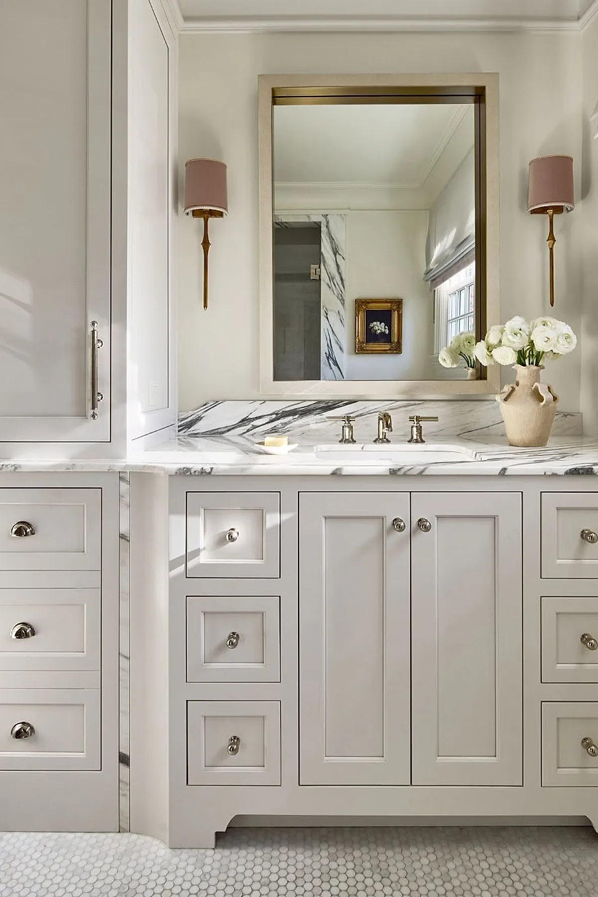 Cream and white bathroom featuring a marble countertop with gray veining, cream cabinetry with silver knobs, brass wall sconces with mauve shades on either side of a large rectangular mirror framed in light wood, and a beige ceramic vase holding white flowers.