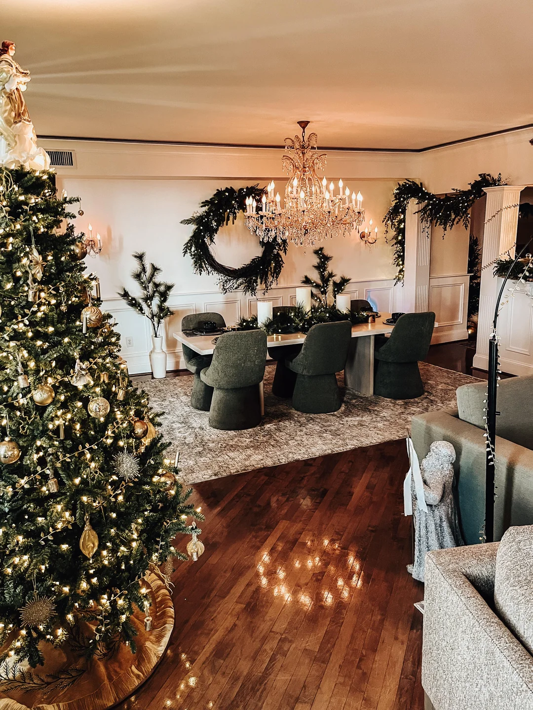 traditional Christmas tree decorated with gold ornaments and warm lights beside a chandelier-lit dining room with greenery garlands and wreaths