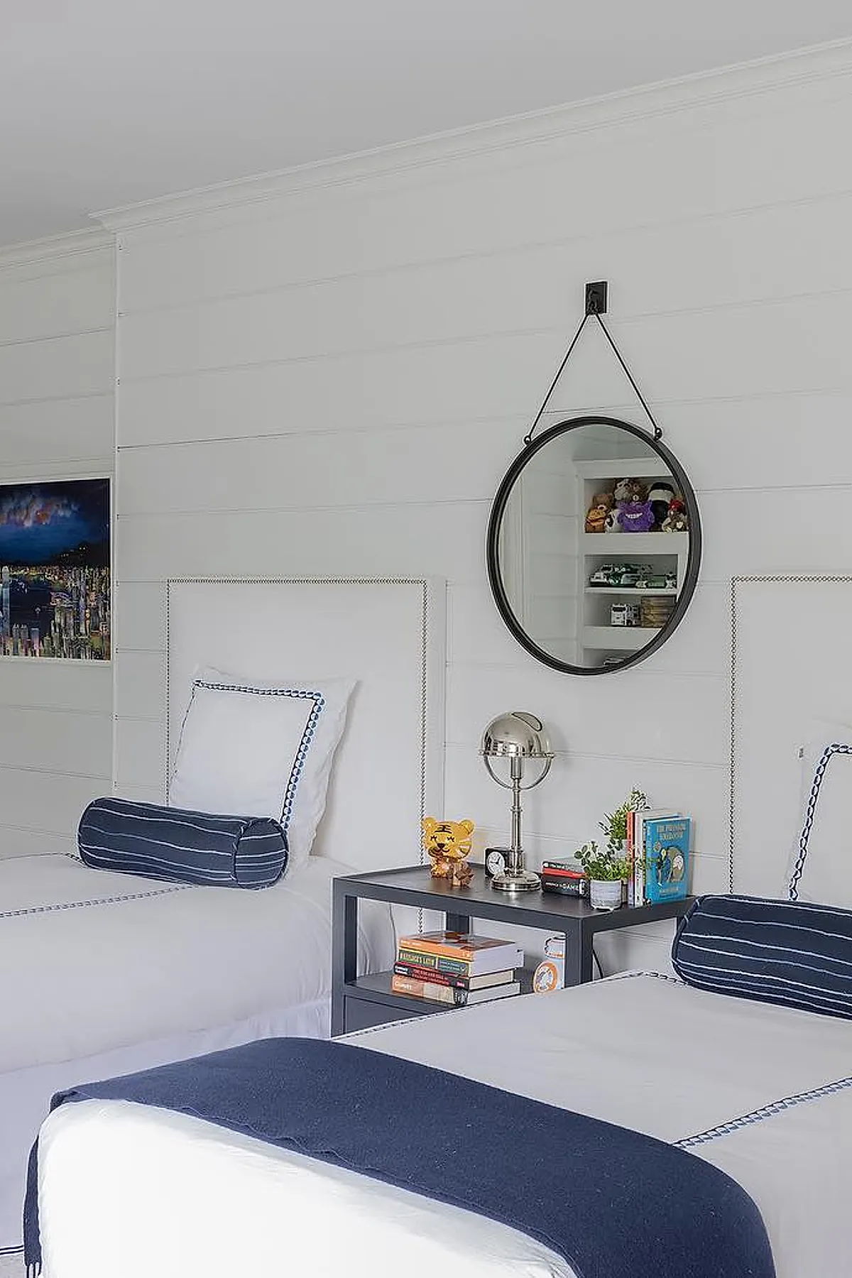 Two twin white beds with navy-trimmed pillows and navy striped bolsters separated by a dark gray nightstand holding books, a silver desk lamp, plant, and toys. A round black-framed mirror hangs on the white shiplap wall above the nightstand.