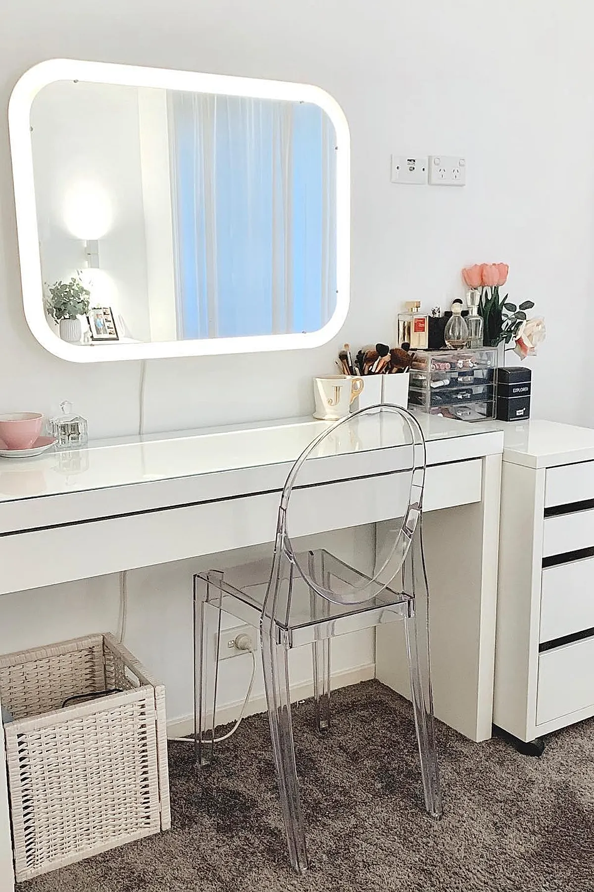 white bedroom vanity with large rectangular LED mirror mounted on wall, clear acrylic chair, white desk with makeup brushes, perfume bottles, pink flowers, wicker basket underneath