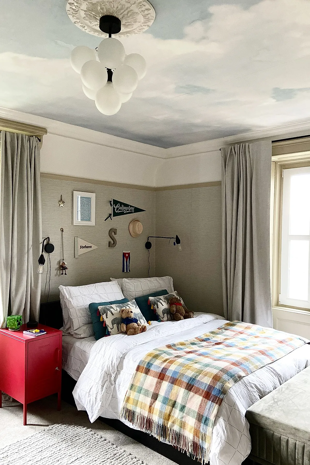 Teen boy bedroom featuring a bed with white grid bedding, multicolored plaid throw blanket, animal print pillows, stuffed teddy bears, beige textured walls decorated with small pennants and framed prints. Ceiling is painted with soft blue sky and white clouds. Curtains are light gray and there is a red bedside cabinet.