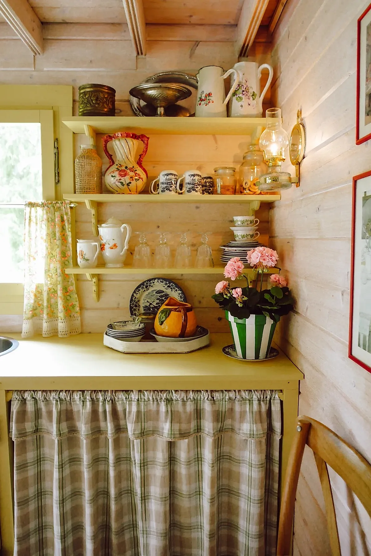 Yellow cottage kitchen featuring light yellow wooden shelves filled with vintage floral pitchers, blue and white mugs, glassware, stacked plates and cups, a striped green and white flower pot with pink flowers on the countertop, cream-colored curtains with floral pattern by a window, wood panel walls, and a warm glowing wall lamp.