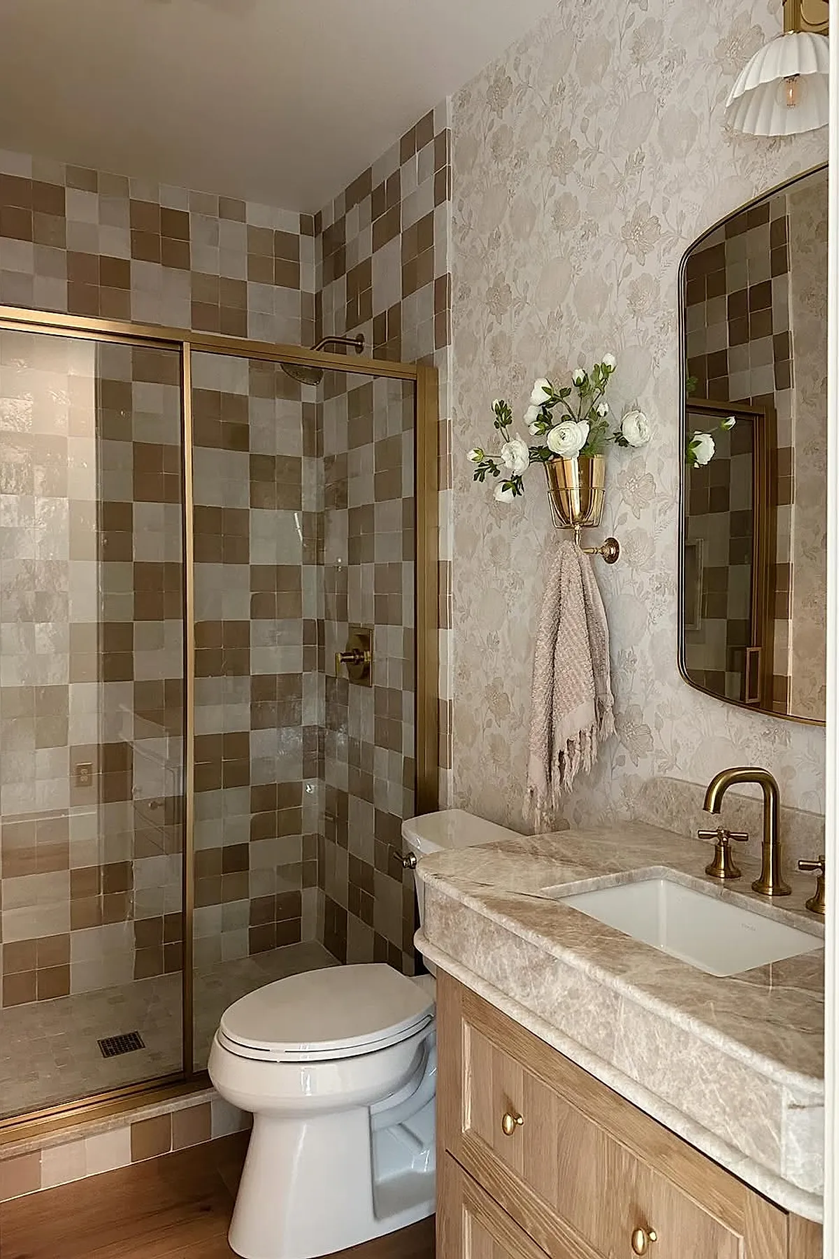 Warm neutral bathroom featuring beige and brown checkered tiles in the shower, brass framed glass shower door, brass faucet and mirror frame, floral wallpaper, wooden vanity with marble countertop, and white toilet.