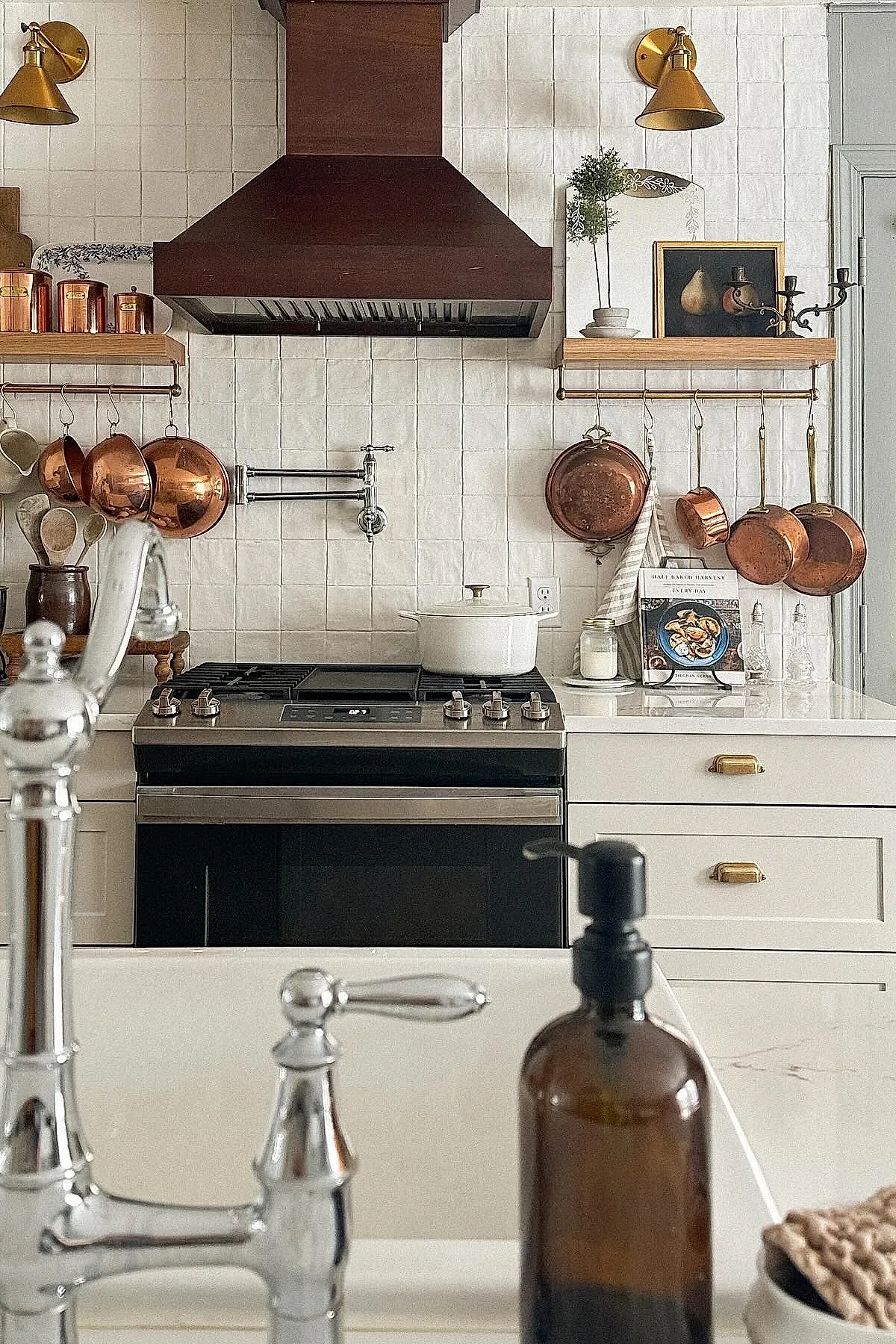 Rustic cottage kitchen backsplash with irregular white square tiles, dark wooden range hood, brass sconces, copper pots and pans hanging from brass rods below wooden shelves holding copper canisters, ceramic utensils in a brown jar, and a white stove with black cooktop.