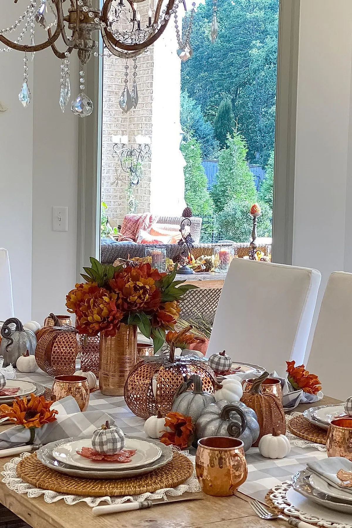 Fall dining table with copper and gray pumpkins, orange flowers in a copper vase, plaid napkins on white plates, wicker chargers, set on a checkered table runner.