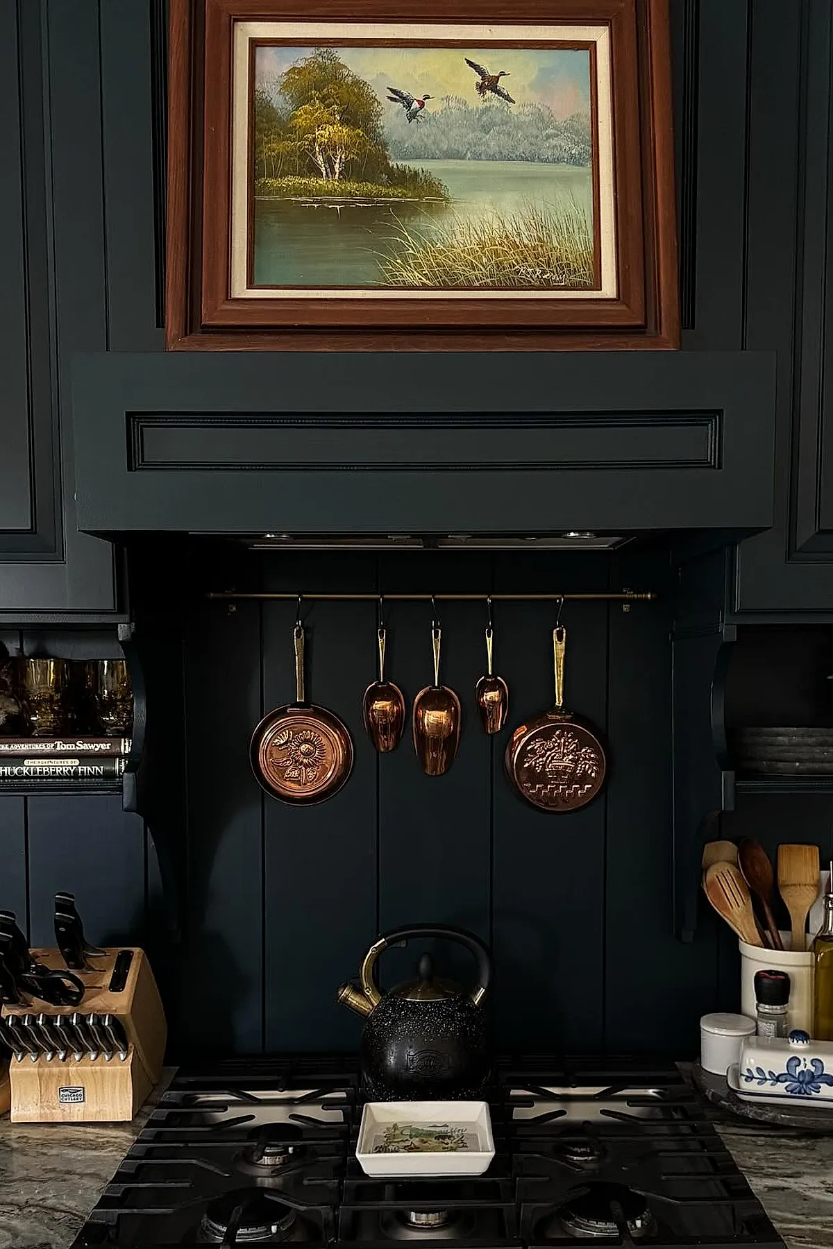 Cottage kitchen backsplash painted dark green or black with copper pans hanging on brass rod, wooden framed landscape painting above stove, knife block on marble countertop, and wooden utensils in white ceramic jar.
