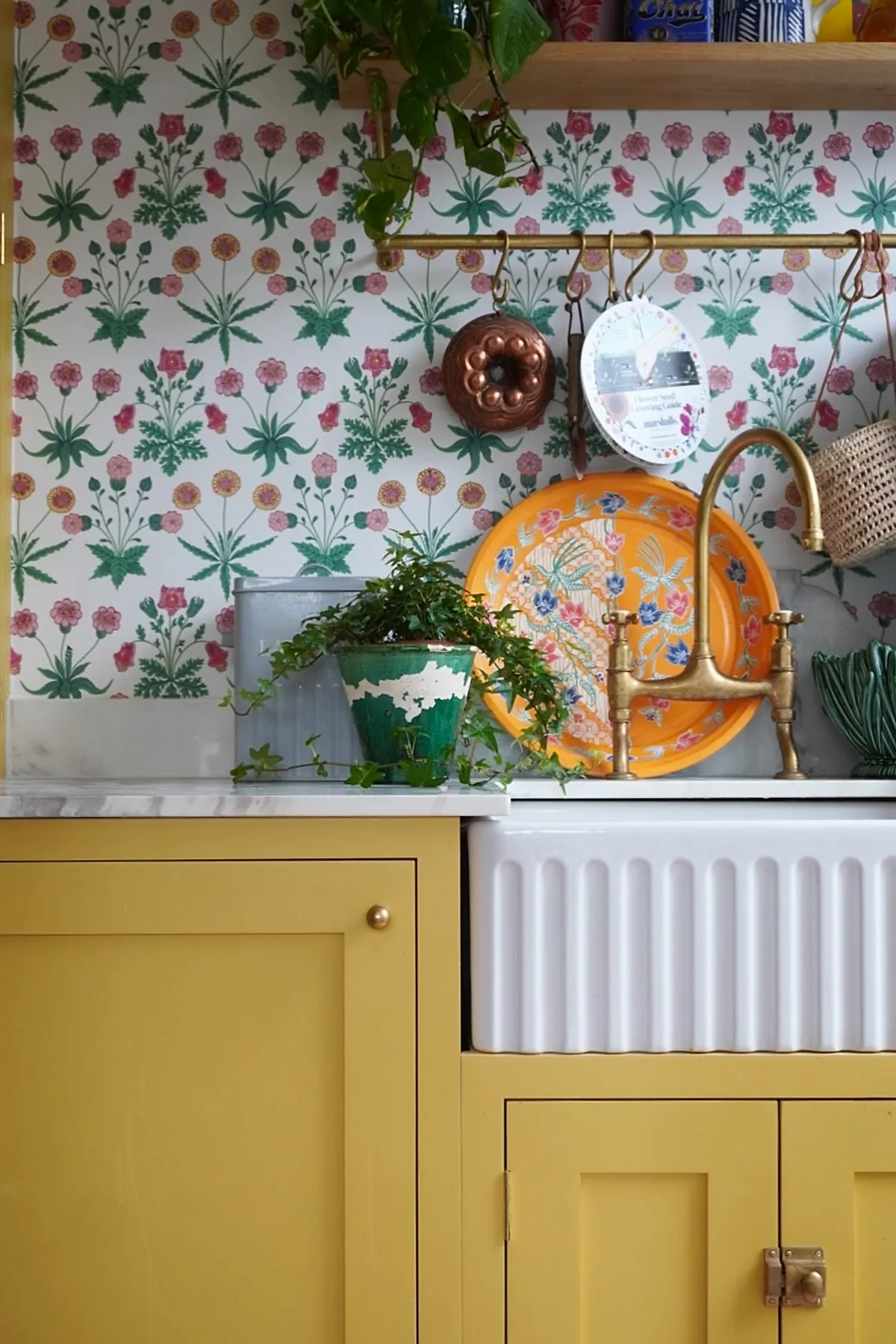 Yellow cottage kitchen with light yellow kitchen walls and cabinetry, white marble countertop, brass faucet over a white farmhouse sink, floral patterned wallpaper in green and pink hues behind the sink area, a vibrant orange floral tray leaning against the backsplash, hanging copper molds and small potted plants adding vintage charm.