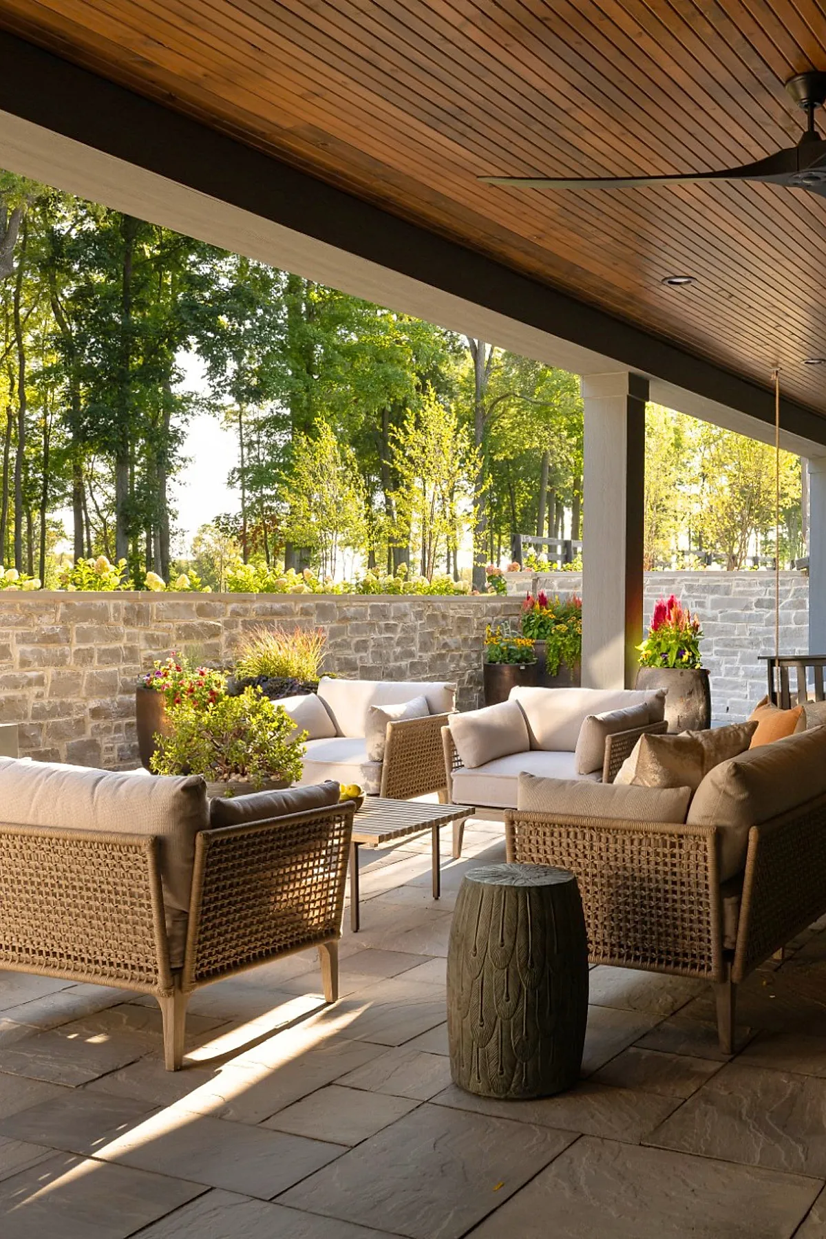 Covered patio attached to house featuring wicker armchairs and sofas with light cushions, stone wall, large ceramic planters with colorful flowers, wooden ceiling with recessed lights, and view of lush green trees.