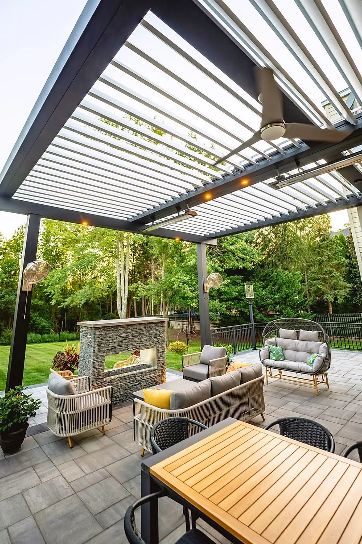 Modern covered patio attached to house with adjustable louvered roof, cushioned wicker seating, stone fireplace, ceiling fan, outdoor dining table, and lush green backyard.