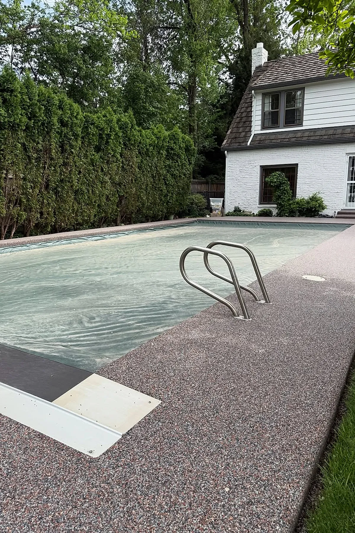 Rectangular swimming pool with a translucent protective cover surrounded by a textured speckled gray pool deck, bordered by tall green hedges and adjacent to a white two-story house with brown roof shingles.