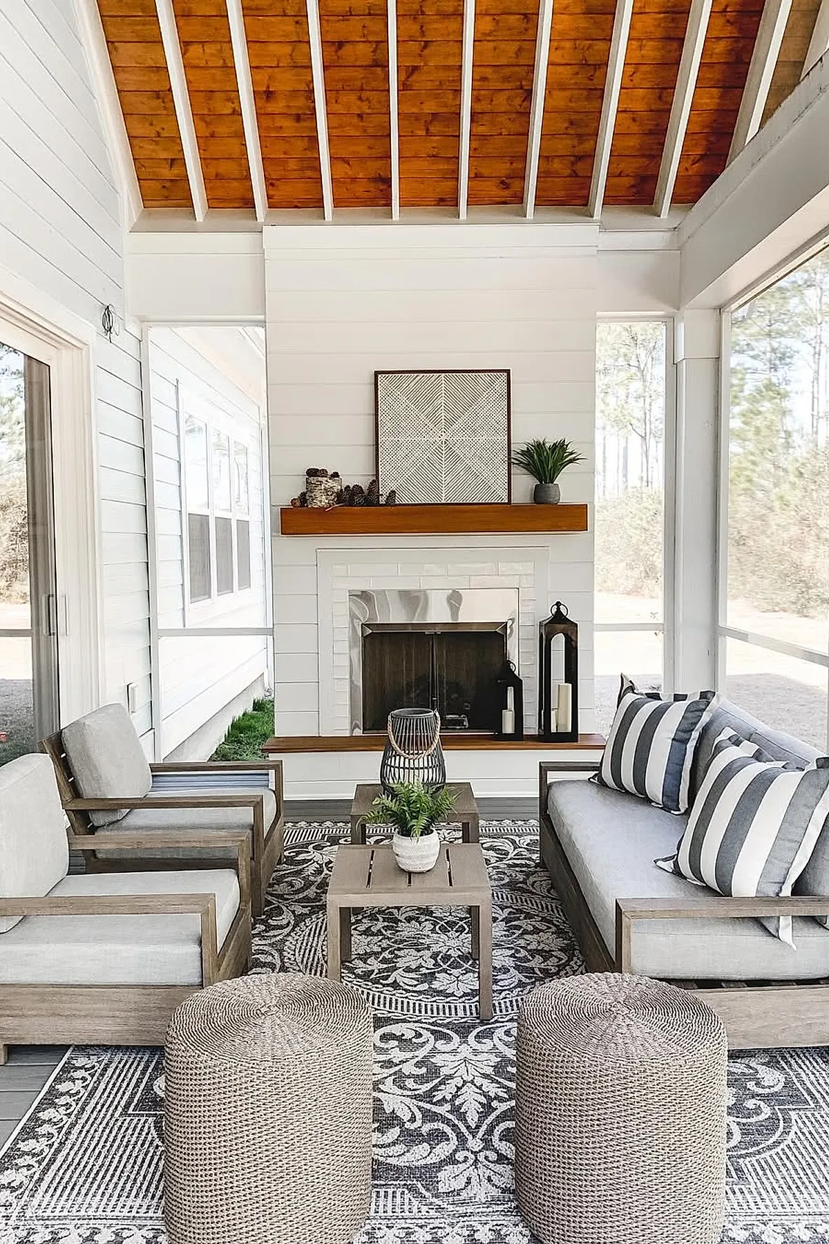 Screened in porch with natural wood vaulted ceiling, white paneled walls, gray cushioned wooden armchairs and sofa with striped pillows, patterned area rug, small coffee tables with decorative plants and lanterns, fireplace with wood mantel