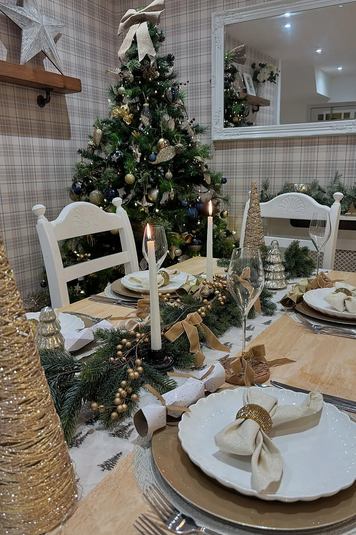 christmas dinner table settings featuring white plates with gold chargers, cream napkins with gold rings, tall white taper candles, gold decorative trees, greenery garland with gold berries on a wooden table in front of a decorated Christmas tree and plaid wallpaper.