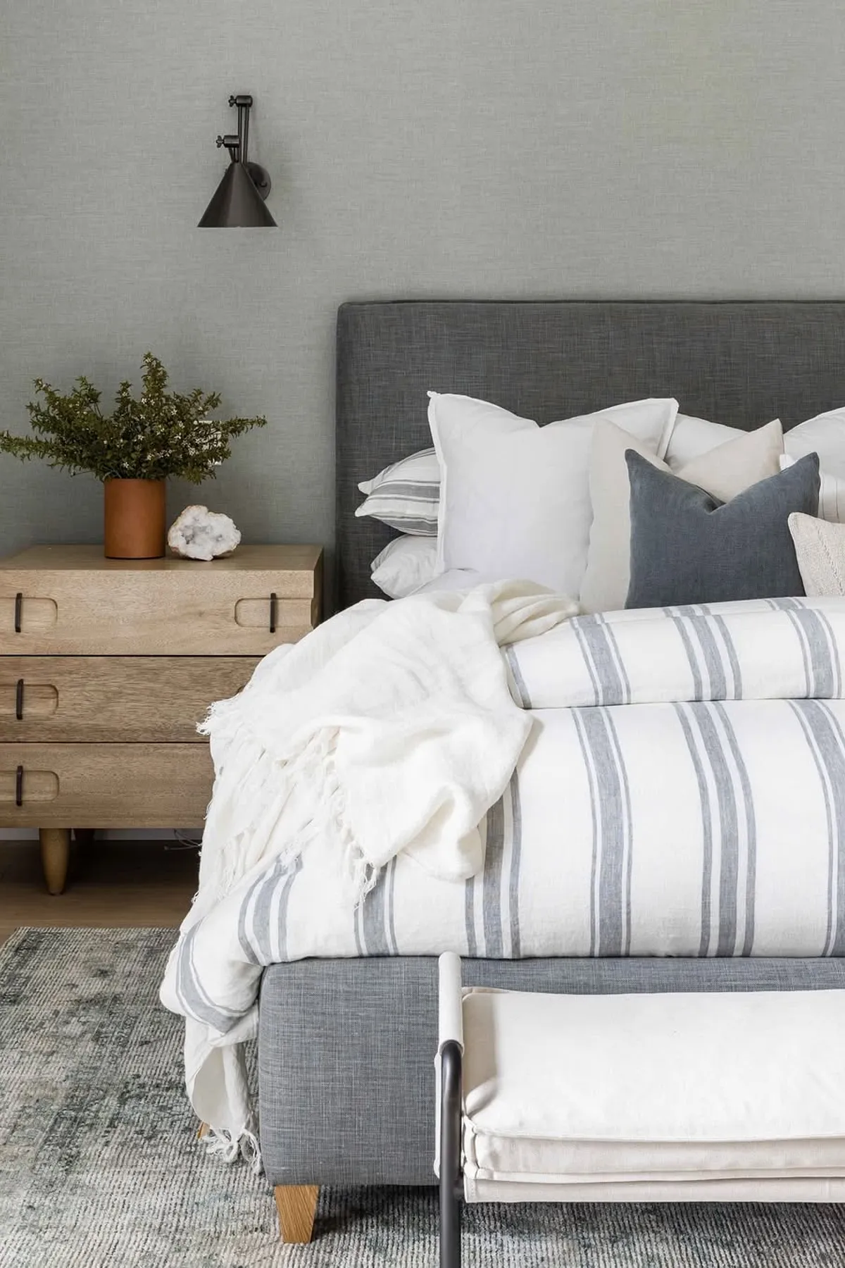 Serene bedroom with striped bedding, gray headboard, wooden nightstand with greenery, modern wall lamp, and textured gray rug.
