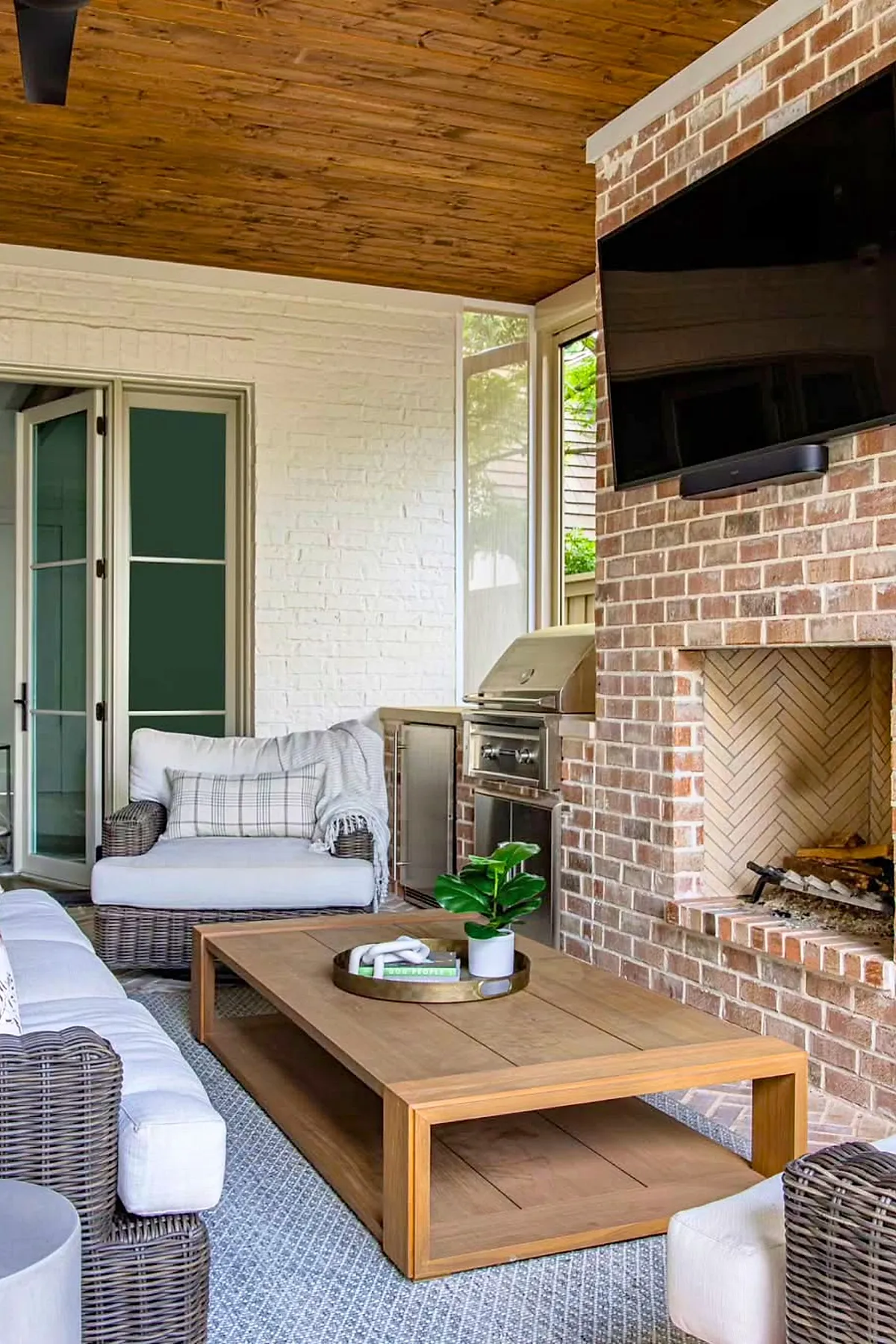 Covered patio attached to house featuring brick fireplace, mounted TV, rattan armchairs and sofa with white cushions, wooden coffee table with decorative tray and plant, wood plank ceiling, and stainless steel built-in outdoor grill.