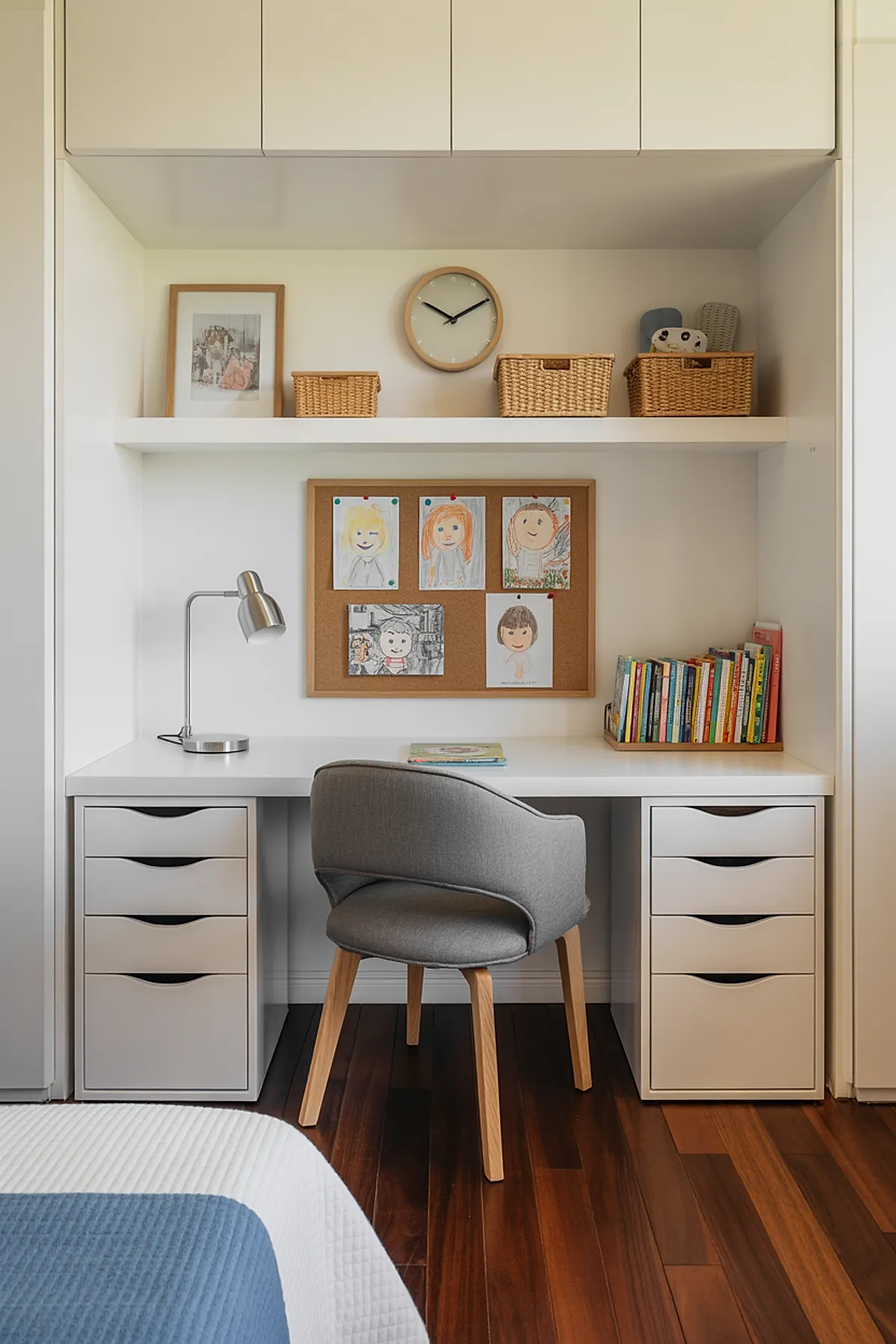 Minimalist kids room with built-in white kids study desk featuring multiple drawers on each side, gray upholstered chair with wooden legs, cork bulletin board displaying children