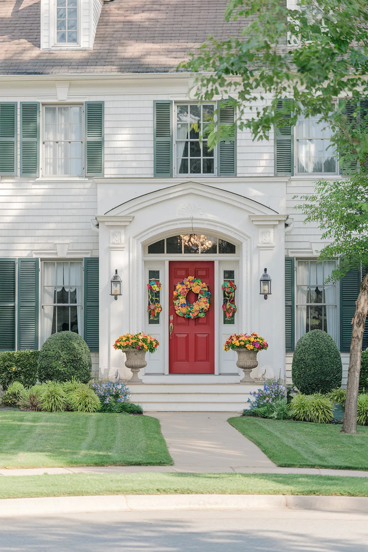 Red front door with a colorful floral wreath and matching planters on a white house.