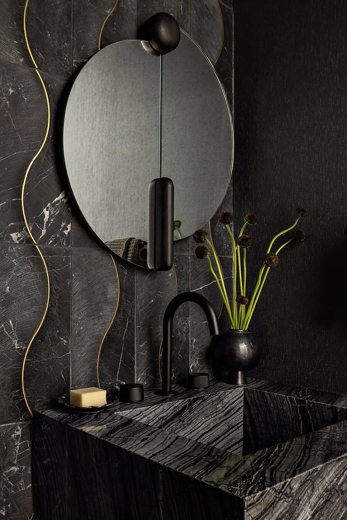 Round bathroom mirror with black metal fixture above a black marble sink featuring striking veining, set against dark textured wall tiles accented with thin gold lines.