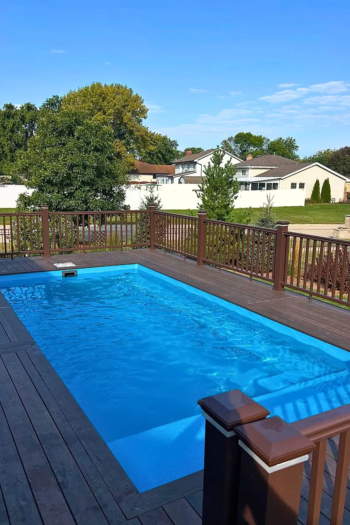 Small rectangular inground pool with bright blue water, surrounded by dark wood decking and brown wooden railings, set in a suburban backyard with green trees and houses visible beyond a white fence under a clear blue sky.