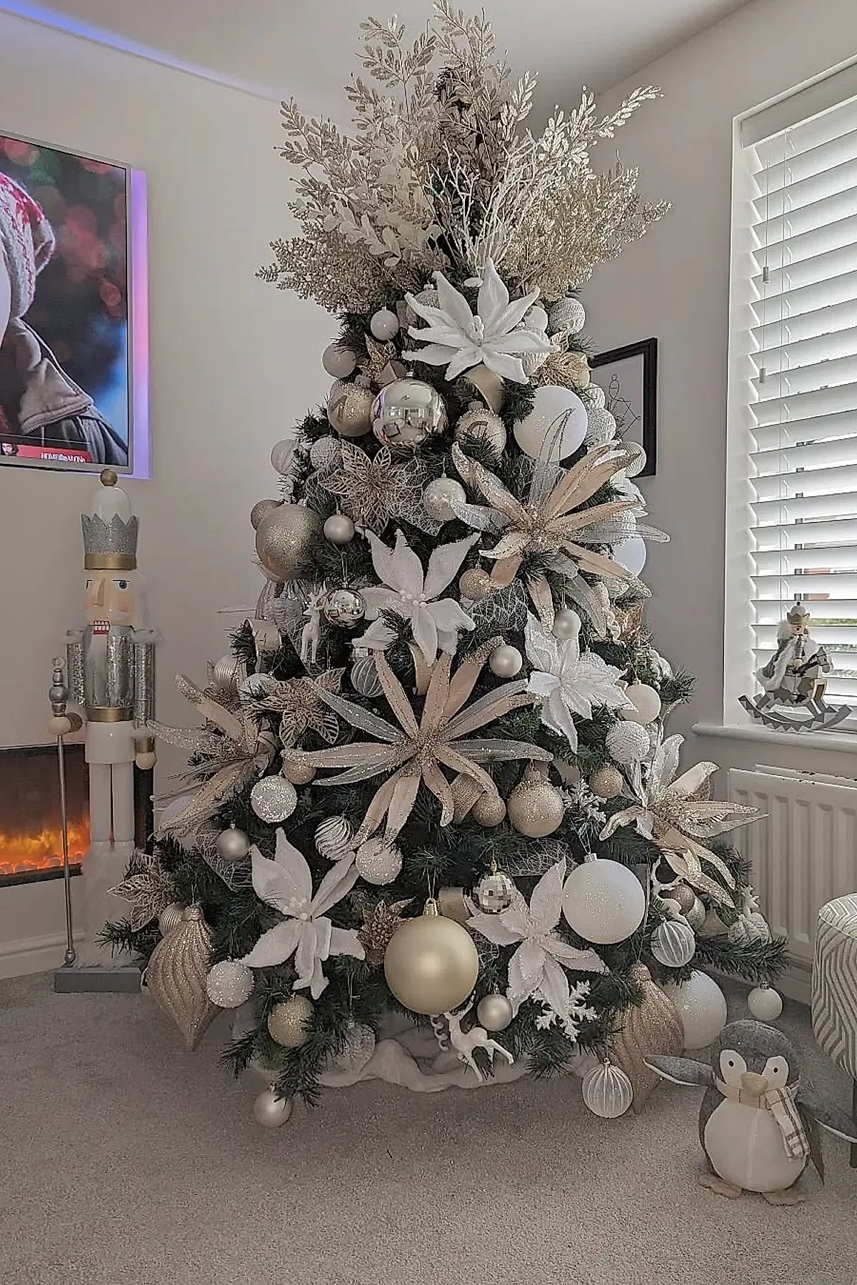Dark green Christmas tree decorated with large white fabric flowers, gold glittered leaves, various sizes of white and gold baubles, set beside a window with blinds and near a nutcracker figure and plush penguin.