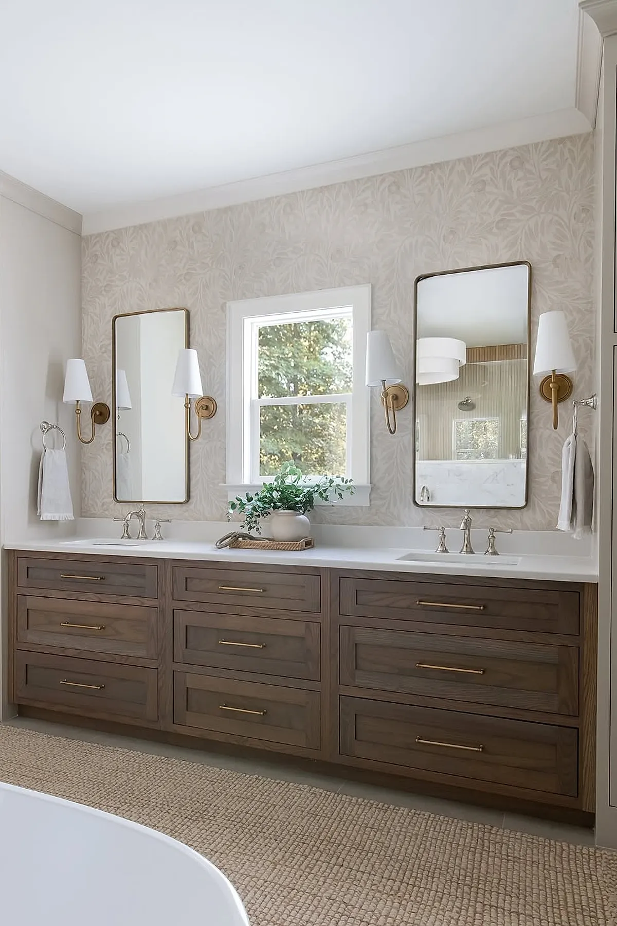 Warm neutral bathroom with a double vanity featuring dark wood drawers with brass handles, two rectangular mirrors with brass frames, wall sconces with white shades on either side of each mirror, light cream wallpaper with subtle leaf pattern, and a window centered between the mirrors.