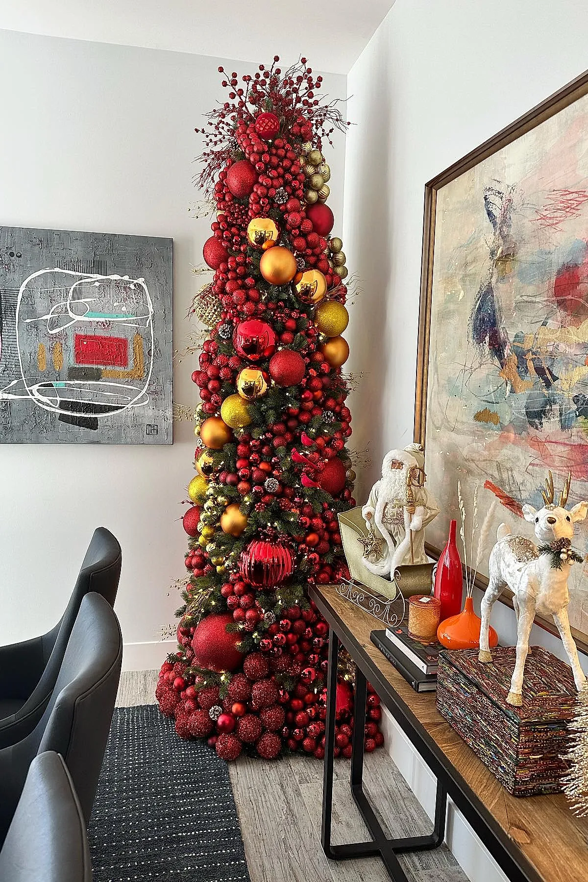 Tall narrow Christmas tree densely decorated with clusters of red berries, large red and gold baubles in living room corner next to side table holding white Santa figure, white reindeer statue, red vase, and abstract wall art