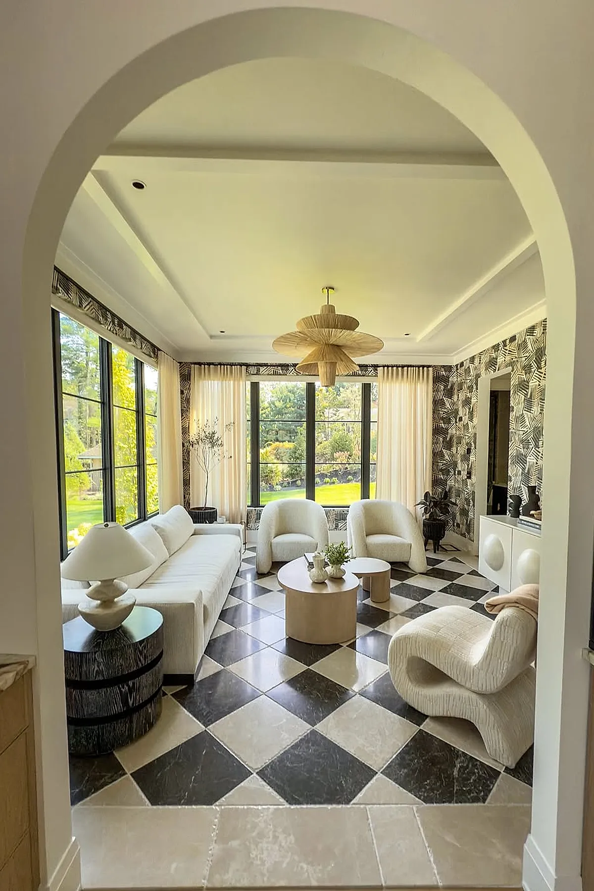 Modern sunroom with black and white checkered tile floor, white curved armchairs and sofa, round light wood coffee tables, large windows with black frames, patterned black and white wallpaper, woven pendant light, viewed through an arched doorway.