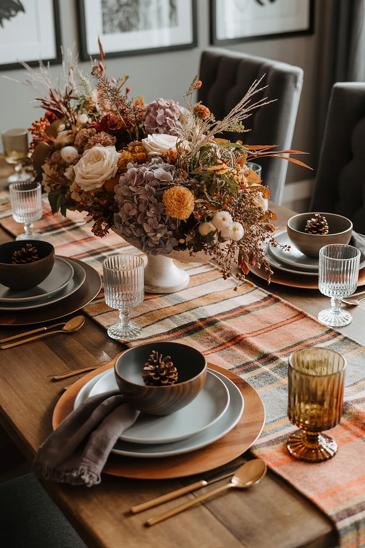 Thanksgiving table set with brown wood surface featuring plaid orange and cream runner, white pedestal vase holding dried flowers in purple, cream, yellow and burnt orange tones; place settings with gray plates, wooden bowls holding pinecones; gold flatware; amber and clear ribbed glasses