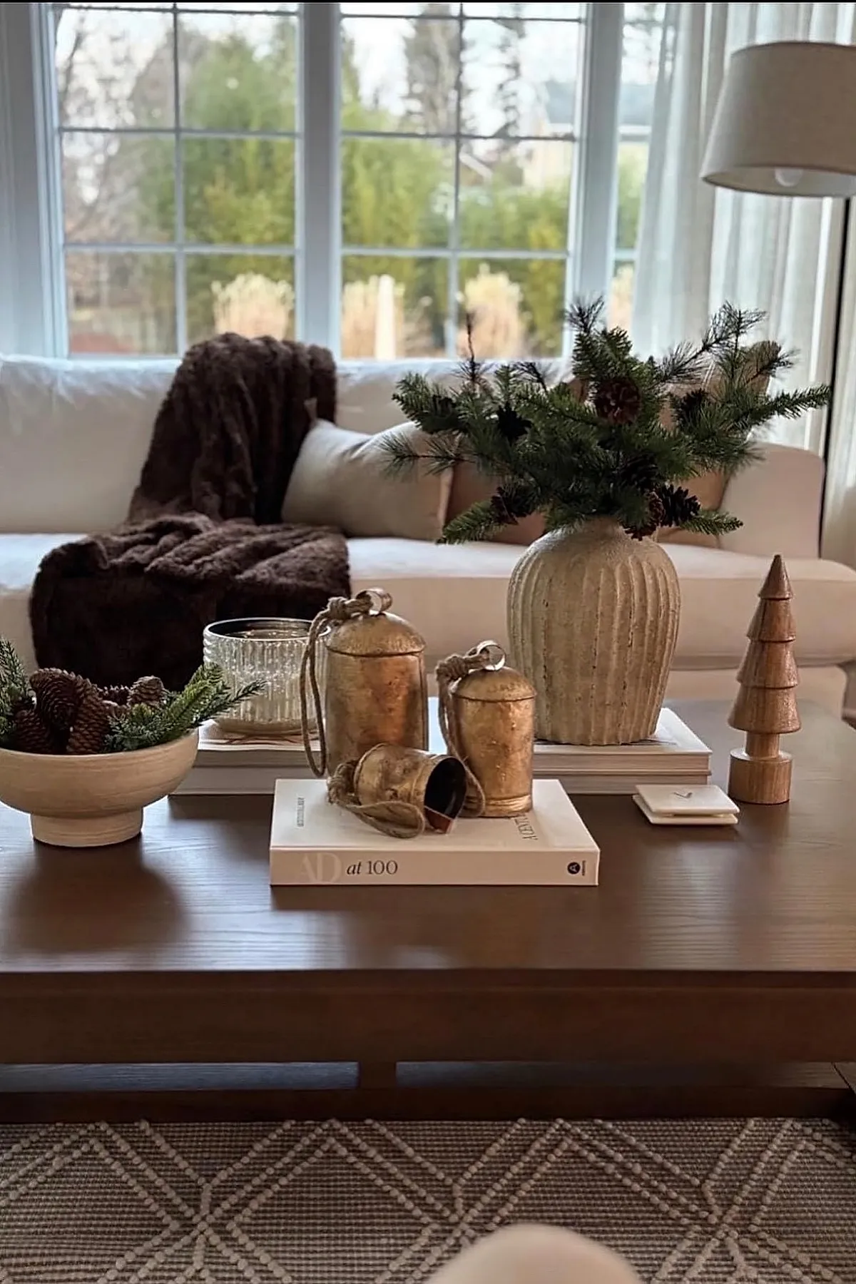 christmas coffee table decor featuring a wooden bowl with pinecones and greenery, three golden bells on a book, a textured ceramic vase holding pine branches with pinecones, and a small wooden tree sculpture on a dark wood coffee table in front of a white sofa with brown throw.