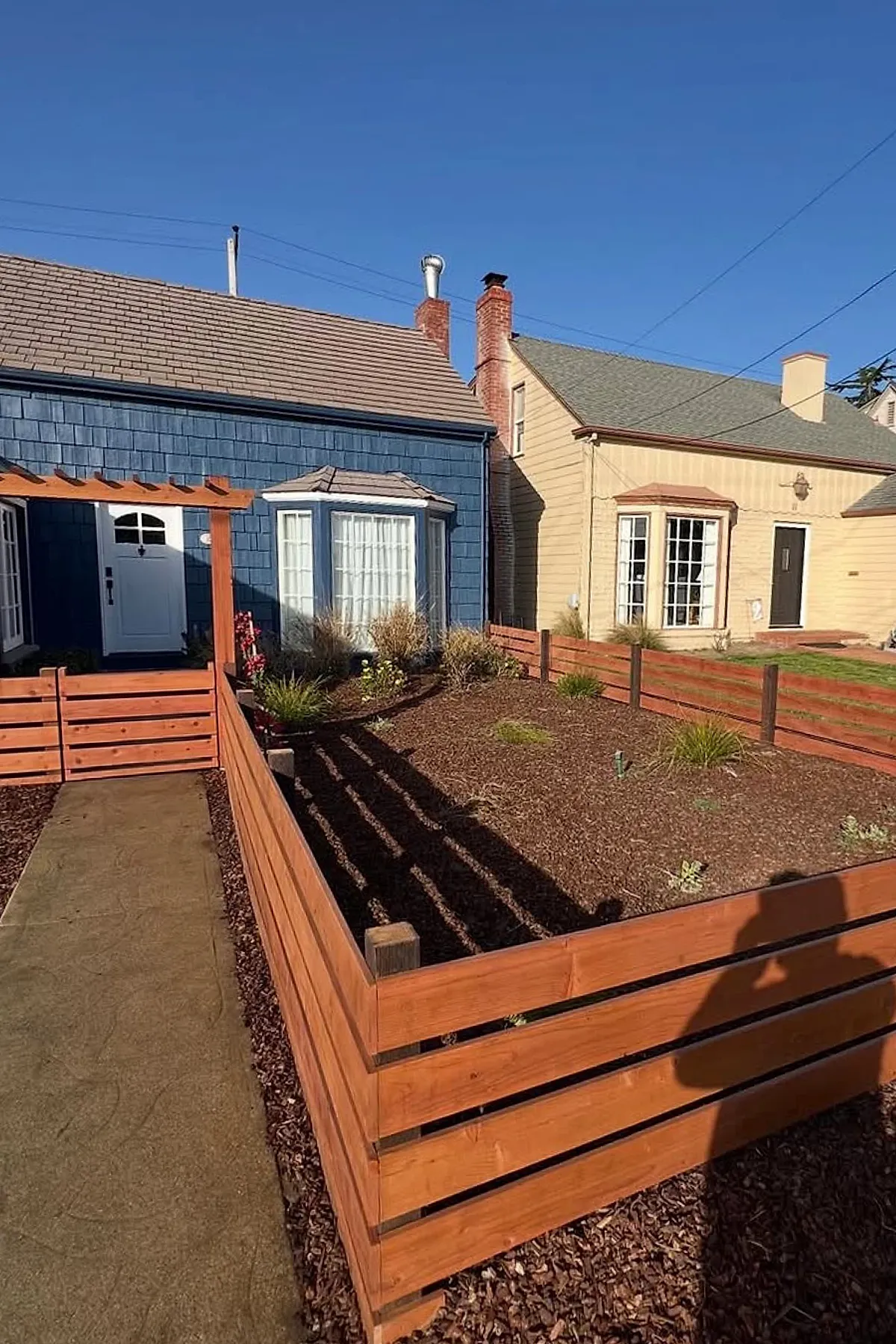 Low front yard fence made of horizontal wooden planks with a matching wooden pergola in front of a blue house with a bay window and white door under clear blue sky.