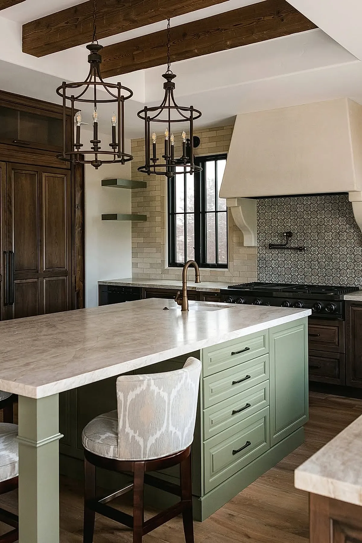 Spanish style kitchen featuring a large marble countertop island painted soft green with black hardware, dark wood cabinets, wrought iron pendant lights, beige brick backsplash, patterned tile behind the stove, and exposed wooden ceiling beams.